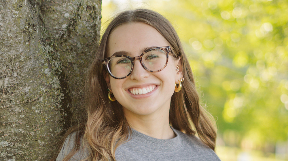 Portrait of a current Gannon Scholar with long light brown hair and glasses, smiling, wearing a gray top and gold hoop earrings, standing outdoors beside a tree with greenery in the background.